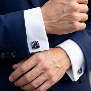 Op de afbeelding: Close-up van vierkante manchetknopen met een blauw en zilver bloemmotief, tegen een witte overhemdmouw en een marineblauw pak. De manchetknopen zijn het middelpunt van de afbeelding.