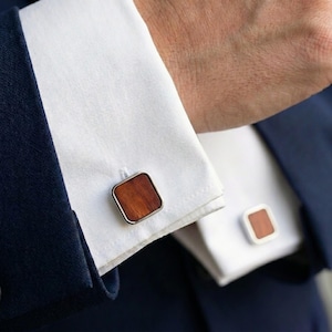Può includere: Primo piano di un paio di gemelli. I gemelli hanno un centro quadrato in legno con un bordo in metallo color argento. I gemelli sono su una camicia bianca con una giacca blu navy.