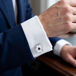 Puede incluir: Gemelos plateados con el diseño de la carta del As de Picas. Los gemelos rectangulares tienen un fondo blanco con símbolos de cartas negras y rojas. Están en un puño de camisa blanco, combinados con un traje azul marino.