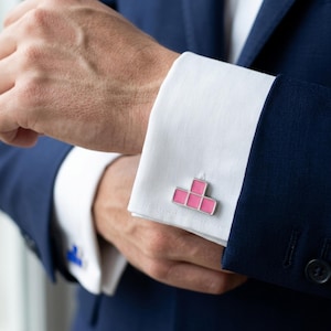 May include: Silver cufflinks with a pink geometric design are displayed on a white dress shirt cuff. The cufflinks are a bright pink color with a silver border. The person is wearing a navy blue suit.