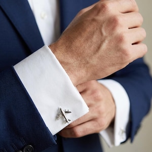 Puede incluir: Gemelos plateados con forma de avión en un puño de camisa blanca. Los gemelos presentan alas, fuselaje y cola. La camisa se combina con una chaqueta de traje azul marino.