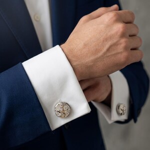 May include: A pair of silver cufflinks featuring intricate watch movement details. The round cufflinks are set on white shirt cuffs, contrasted by a navy blue suit jacket. The watch mechanism is visible.