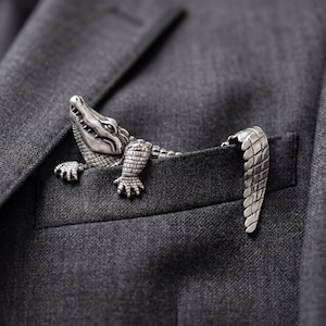 Könnte beinhalten: Ein silberner Alligator-Reversnadel, mit detaillierten Schuppen und Krallen, ragt aus einer grauen Anzugtasche. Der Kopf und die Vorderbeine des Alligators sind sichtbar, während sein Schwanz aus der Tasche hängt.