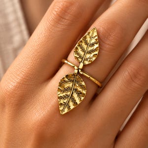 May include: A gold-toned ring featuring two detailed leaf-shaped charms. The leaves are positioned vertically, one pointing upward and one pointing downward, connected by a thin, polished gold band that wraps around the finger.