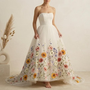May include: A strapless white wedding dress with a floral embroidered skirt. The dress features a fitted bodice and a full, flowing skirt adorned with colorful embroidered flowers, including orange, red, and purple blooms. The dress has a long train.