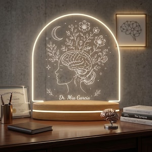 May include: A decorative night light with a wooden base and a glowing arched acrylic panel. The panel features a line drawing of a woman's profile with a brain and floral elements, and the text "Dr. Mia Garcia". A framed brain illustration is on the wall.