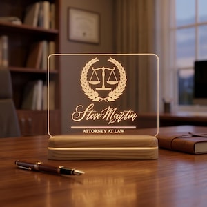 May include: A personalized desk lamp with a wooden base and a clear acrylic panel. The panel features a gold-colored scales of justice emblem, the name "Steve Martin", and the text "ATTORNEY AT LAW". The lamp is on a wooden desk.