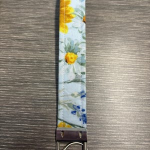 May include: A floral key fob featuring a light blue background with yellow and blue flower patterns. The fabric is attached to a silver-colored metal key ring and hardware.