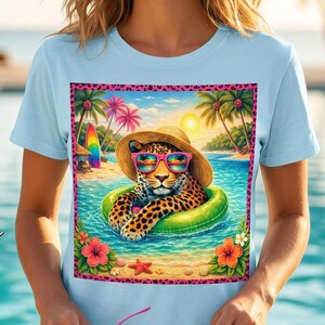 May include: Pale blue t-shirt with a colourful graphic of a leopard wearing sunglasses and a sun hat, floating on an inner tube in a tropical beach scene. The design includes palm trees, ocean, and a vibrant border. The woman is holding pink sunglasses.