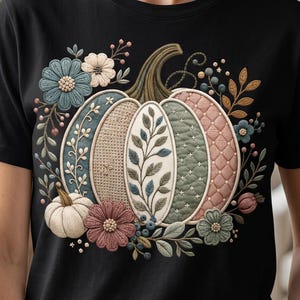May include: A black short-sleeved t-shirt featuring a central graphic of a large pumpkin decorated with various textures and patterns, surrounded by a wreath of colourful flowers, leaves, and berries in muted tones of blue, pink, and green.