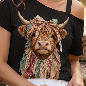 May include: A black short-sleeved t-shirt featuring a graphic print of a brown Highland cow wearing a multi-coloured floral patterned fabric bow and a fringed neck scarf. The shirt has an asymmetrical off-the-shoulder neckline design.