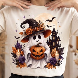 May include: A cream-coloured t-shirt featuring a graphic of a ghost wearing a witch hat and holding a jack-o'-lantern. The design includes a haunted house, bats, a full moon, lit candles, and autumn leaves in shades of orange and purple.