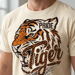 May include: Cream-colored t-shirt featuring a detailed tiger head graphic with the words "Pride Tiger". The tiger design is in shades of orange and black, with a paw print at the bottom.