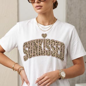 以下が含まれることがあります： ヒョウ柄のデザインで「SUNKISSED」の文字がプリントされた白いTシャツ。その上にハートマークがあります。モデルはサングラス、ゴールドのジュエリー、腕時計を着用しています。背景はニュートラルです。