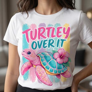 Op de afbeelding: Wit T-shirt met een roze zeeschildpad met een turquoise schild en een roze hibiscusbloem. De tekst "Turtley Over It" staat in roze en turquoise op de voorkant.