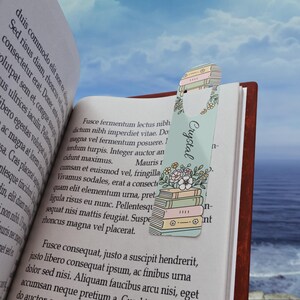 Op de afbeelding: Een lichtblauw bladwijzer met de naam "Crystal" in sierlijke letters, versierd met een stapel boeken en bloemenillustraties. De bladwijzer zit in een open boek met tekst op de pagina's.