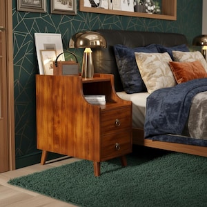 May include: A mid-century modern style wooden nightstand with two drawers and a small open shelf. The nightstand has a warm wood grain finish and is set on angled legs. A gold-toned lamp and a small speaker sit on top.