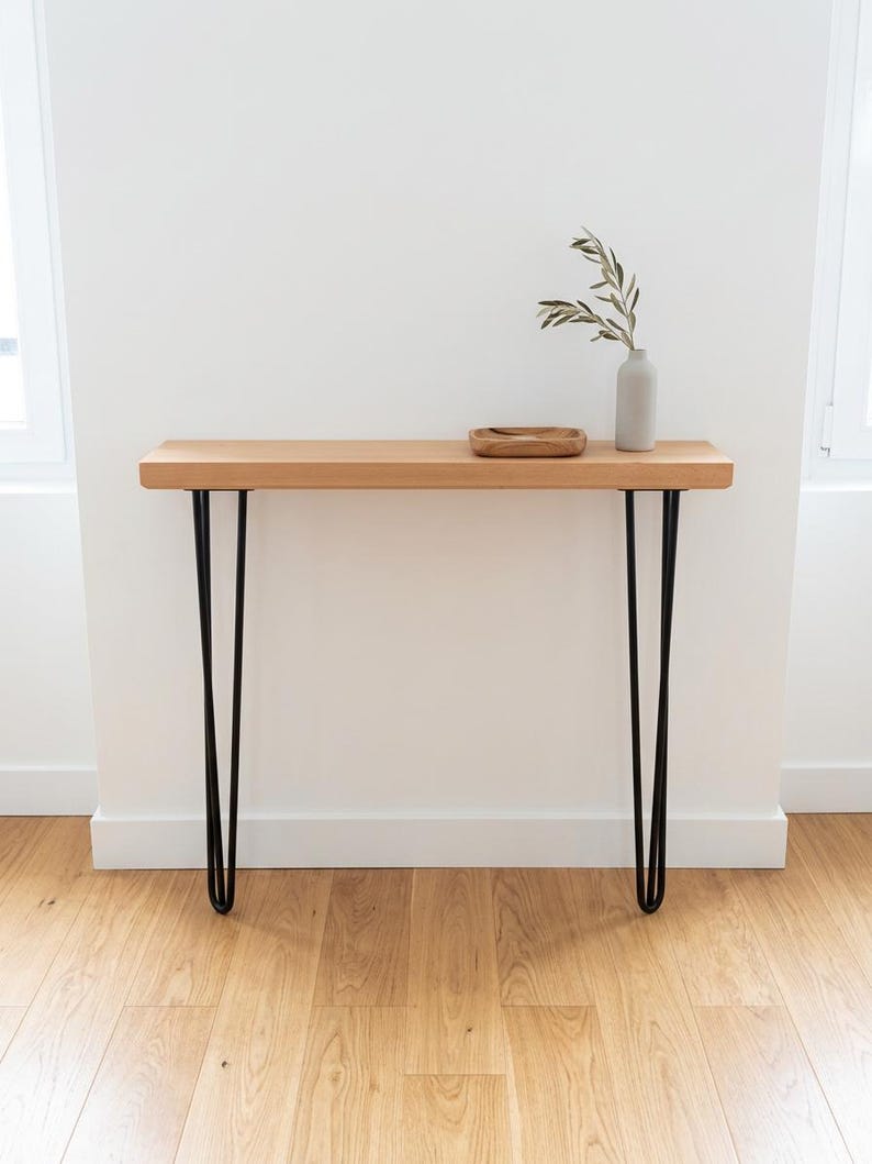 Peut inclure: Une console en bois clair avec des pieds en &eacute;pingle noirs. La table a un plateau rectangulaire et est d&eacute;cor&eacute;e d'un petit plateau en bois et d'un vase gris avec de la verdure. La table mesure environ 122 cm de long.