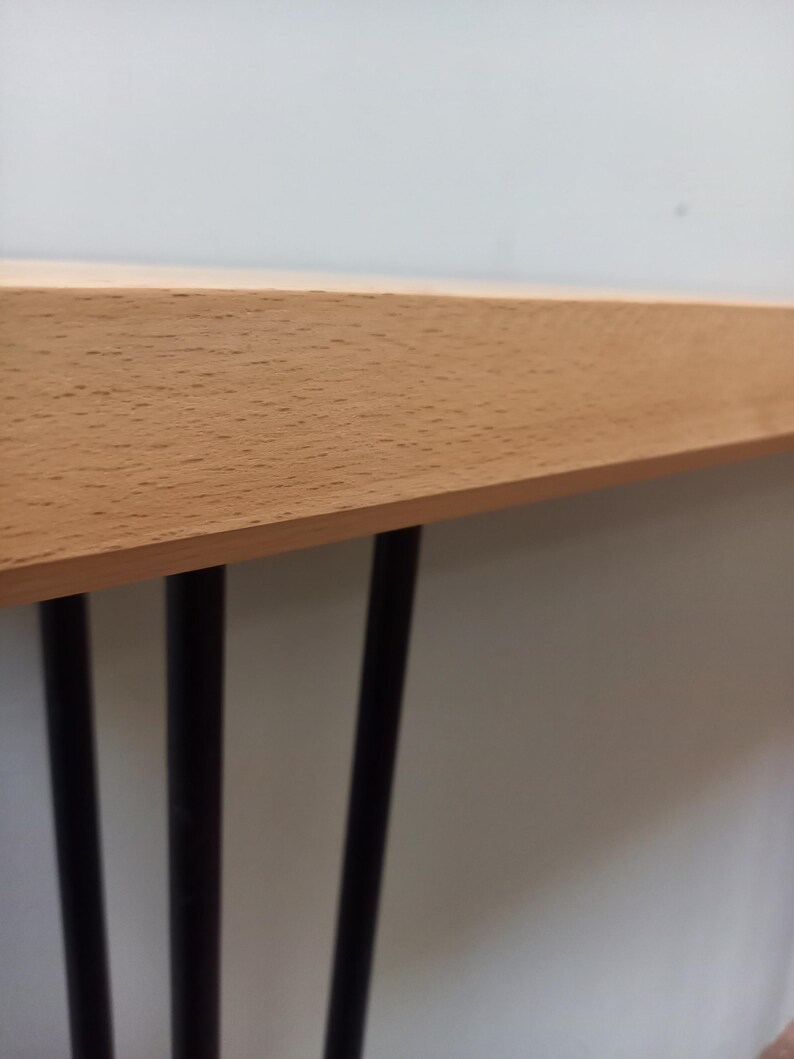 Peut inclure: Une table en bois clair avec trois pieds en m&eacute;tal noir. Le plateau a une finition en grain de bois naturel. Les pieds sont fins et cylindriques, soutenant la table.