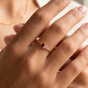 May include: A thin gold ring featuring two small, teardrop-shaped gemstones set side-by-side. One stone is a deep red garnet, and the other is a pale pink stone. The ring has a simple, polished gold band that sits on the finger.