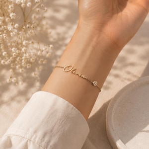 May include: A gold-toned chain bracelet featuring the name Olivia in a cursive script font, accented with a single small, round, clear bezel-set stone positioned along the delicate metal link chain, creating a refined and simple jewelry piece.