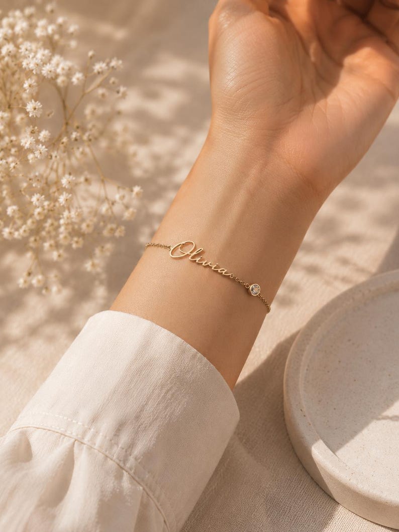 May include: A gold-toned chain bracelet featuring the name Olivia in a cursive script font, accented with a single small, round, clear bezel-set stone positioned along the delicate metal link chain, creating a refined and simple jewelry piece.
