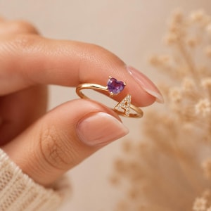 May include: A gold open ring featuring a purple heart-shaped gemstone on one end and a letter A encrusted with small clear stones on the other end. The band is thin and metallic, designed to be adjustable with a gap between the two decorative ends.