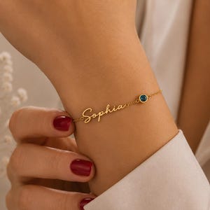 May include: A gold-toned chain bracelet featuring the name Sophia in a cursive script font. The bracelet includes a single small, round, dark blue gemstone set in a gold-toned bezel, positioned along the delicate chain next to the nameplate.