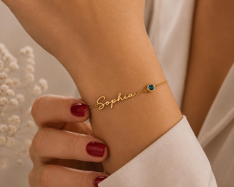 May include: A gold-toned chain bracelet featuring the name Sophia in a cursive script font. The bracelet includes a single small, round, dark blue gemstone set in a gold-toned bezel, positioned along the delicate chain next to the nameplate.