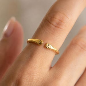 Könnte beinhalten: Ein goldfarbener offener Ring mit zwei abgerundeten Enden, die jeweils mit einem schwarzen Buchstaben graviert sind. Ein Ende zeigt den Buchstaben S und das andere Ende den Buchstaben Z. Der Ring hat eine glatte, polierte Oberfläche und ein offenes Design.
