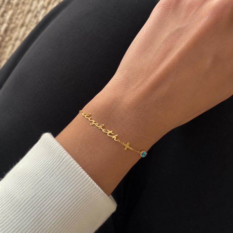 May include: A gold-toned chain bracelet featuring the name Elizabeth in cursive script, a small cross charm, and a single round blue gemstone accent. The delicate metal chain connects these elements, creating a personalized piece of jewelry.