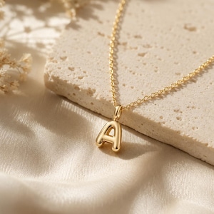 May include: A gold-toned necklace featuring a polished, puffed letter A pendant suspended from a delicate cable chain. The pendant has a smooth, rounded surface and a reflective finish, showcasing a simple, clean design with a classic link chain.