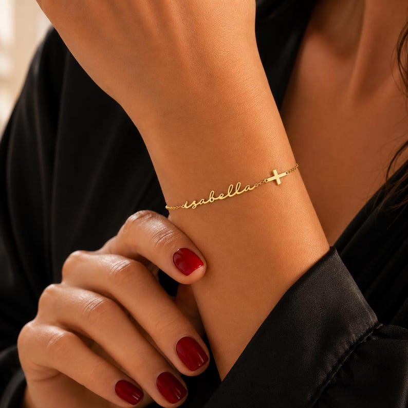 May include: A gold-toned chain bracelet featuring the name Isabella written in a cursive script font, followed by a small, simple cross charm. The delicate metal chain connects to both ends of the nameplate and the cross, creating a continuous link.