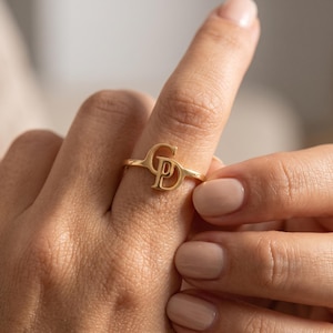 May include: A gold-toned ring featuring a polished finish and a central design composed of the interlocking capital letters G and P. The band is thin and circular, showcasing a metallic luster that reflects light across the surface of the letters.