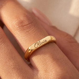 Può includere: Un anello color oro lucido caratterizzato da una superficie superiore piatta e rettangolare. Il nome Olivia è inciso in un carattere corsivo al centro della superficie piatta. L'anello ha una finitura liscia e arrotondata e un aspetto metallico.