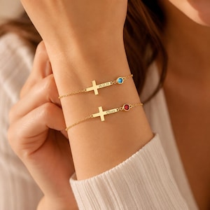 May include: Two gold-tone chain bracelets featuring a horizontal cross charm. One bracelet is engraved with the date 19.12.25 and a blue stone, while the other is engraved with the name Clare and a red stone, both set in a delicate metal link chain.