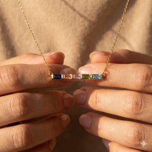 Peut inclure: Un collier en or avec un pendentif en forme de barre orné de cinq pierres rectangulaires bleues, roses, violettes, vertes et bleu clair. Le collier est tenu par une personne.