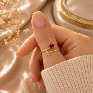 Puede incluir: Un anillo abierto de tono dorado que presenta una pequeña piedra preciosa roja en forma de corazón en un extremo y una letra A mayúscula incrustada con diminutas piedras claras en el otro. La banda es fina y pulida, con un diseño ajustable.