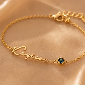 May include: A gold-toned chain bracelet featuring the name Kaylee written in a cursive script font. The bracelet includes a single round, dark blue gemstone set in a gold-toned bezel, a delicate link chain, and a lobster claw clasp closure.