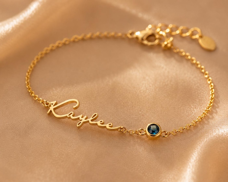 May include: A gold-toned chain bracelet featuring the name Kaylee written in a cursive script font. The bracelet includes a single round, dark blue gemstone set in a gold-toned bezel, a delicate link chain, and a lobster claw clasp closure.