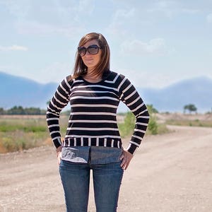 May include: A woman wearing a black and white striped sweater, blue jeans, and brown ankle boots stands on a dirt road. She is wearing sunglasses and has her hands in her pockets.
