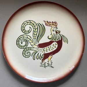 May include: Round ceramic plate with a rooster design. The plate has a cream-colored center with a brown rim. The rooster is depicted in shades of red, yellow, and green, with a detailed tail and comb.