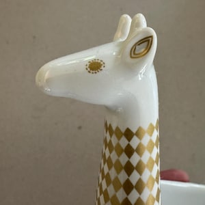 May include: A white ceramic giraffe figurine head and neck featuring a gold diamond pattern along the neck, a gold dotted eye detail, and gold-lined ears. The surface is smooth and glossy, showcasing a stylised representation of a giraffe.