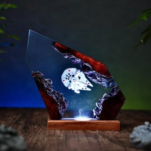 Peut inclure: Une œuvre d'art en résine unique représentant un modèle blanc du Faucon Millenium sur un fond bleu étoilé. L'œuvre est enfermée dans un bloc de résine clair et angulaire avec des accents de bois foncé et de roches, monté sur une base en bois.