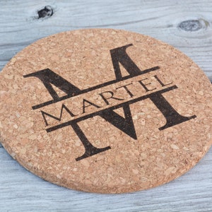 May include: A round cork coaster with a black and white monogram design. The letter "M" is split in half with the name "MARTEL" printed in black across the center.