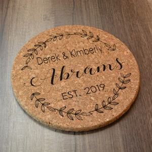 May include: Cork trivet with a black engraved design featuring a wreath of leaves, the text "Derek & Kimberly Abrams EST. 2019", and a decorative script font.
