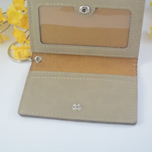 May include: A beige leather card holder with a clear ID window and a metal key ring. The holder has a brown suede lining and a snap closure.