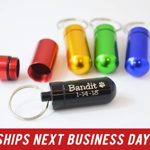 May include: Five small metal pill containers with key rings. The containers are red, yellow, green, blue, and black. The black container has the text "Bandit 1-14-18" engraved on it.