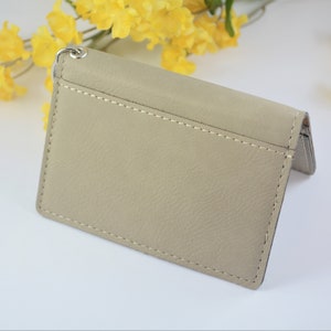May include: A beige leather wallet with a silver chain attached. The wallet is closed and has a stitched seam around the edges.