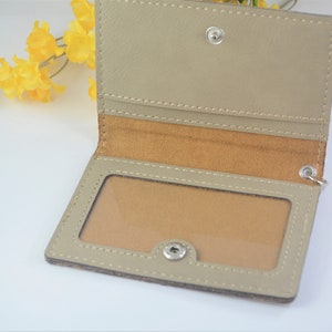 May include: A beige leather card holder with a clear plastic window for displaying an ID card. The holder has a brown suede lining and a silver snap closure.
